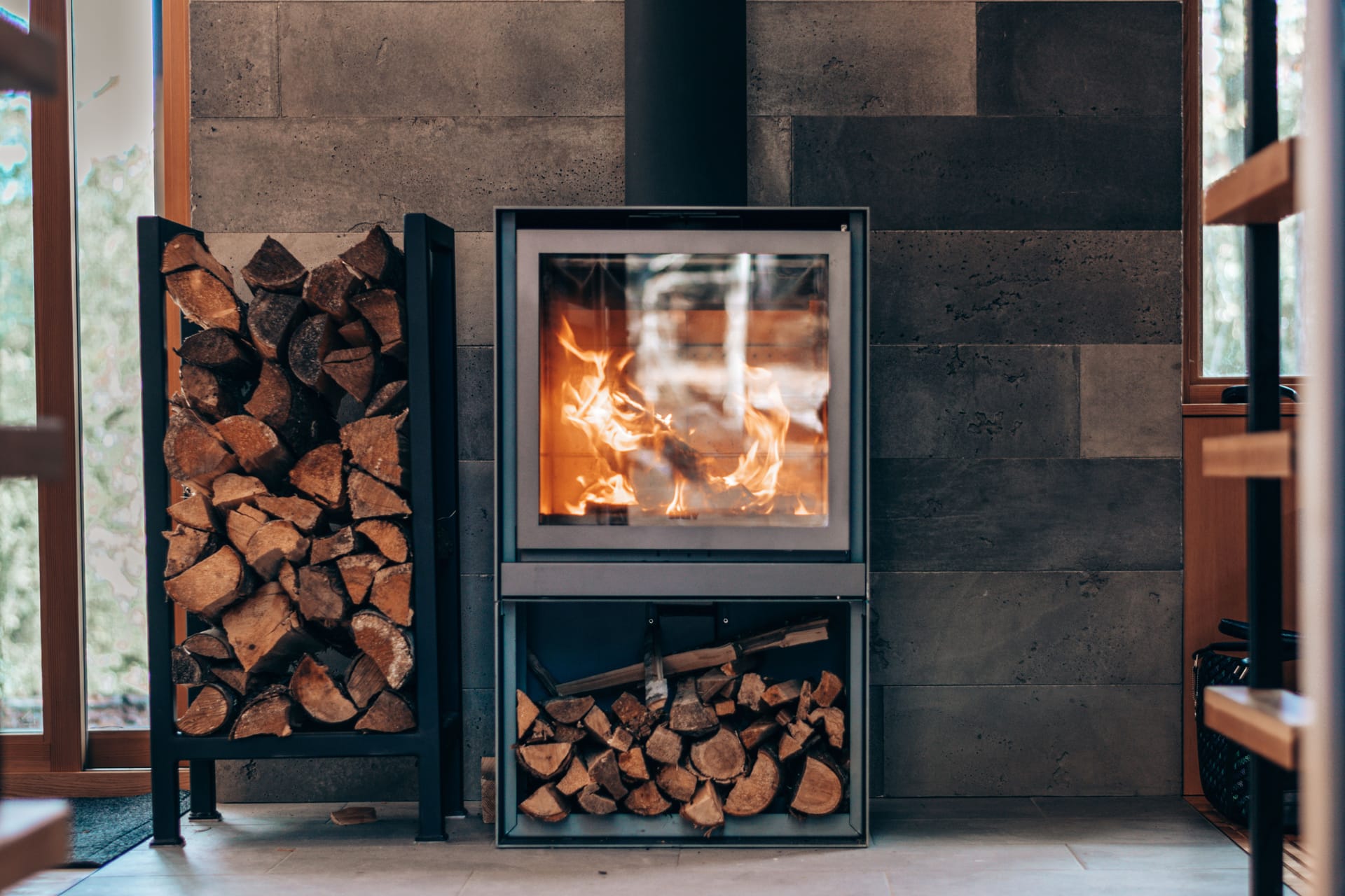 A modern wood-burning stove alight, logs stored beside it